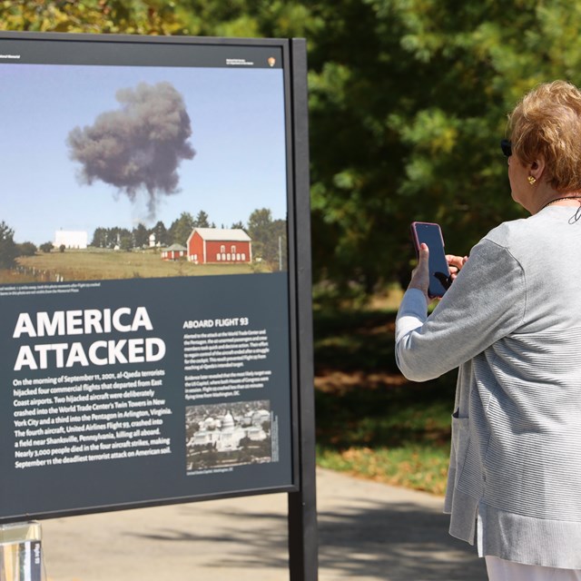 A woman uses her phone at the memorial plaza near an informational panel 