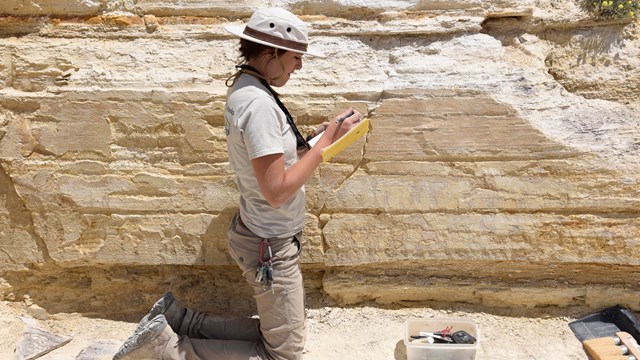 A person in a tan uniform kneeling on rock and writing in a notebook.