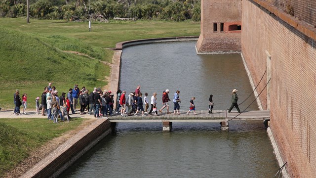 A ranger leading a group over a wooden bridge across a moat. 