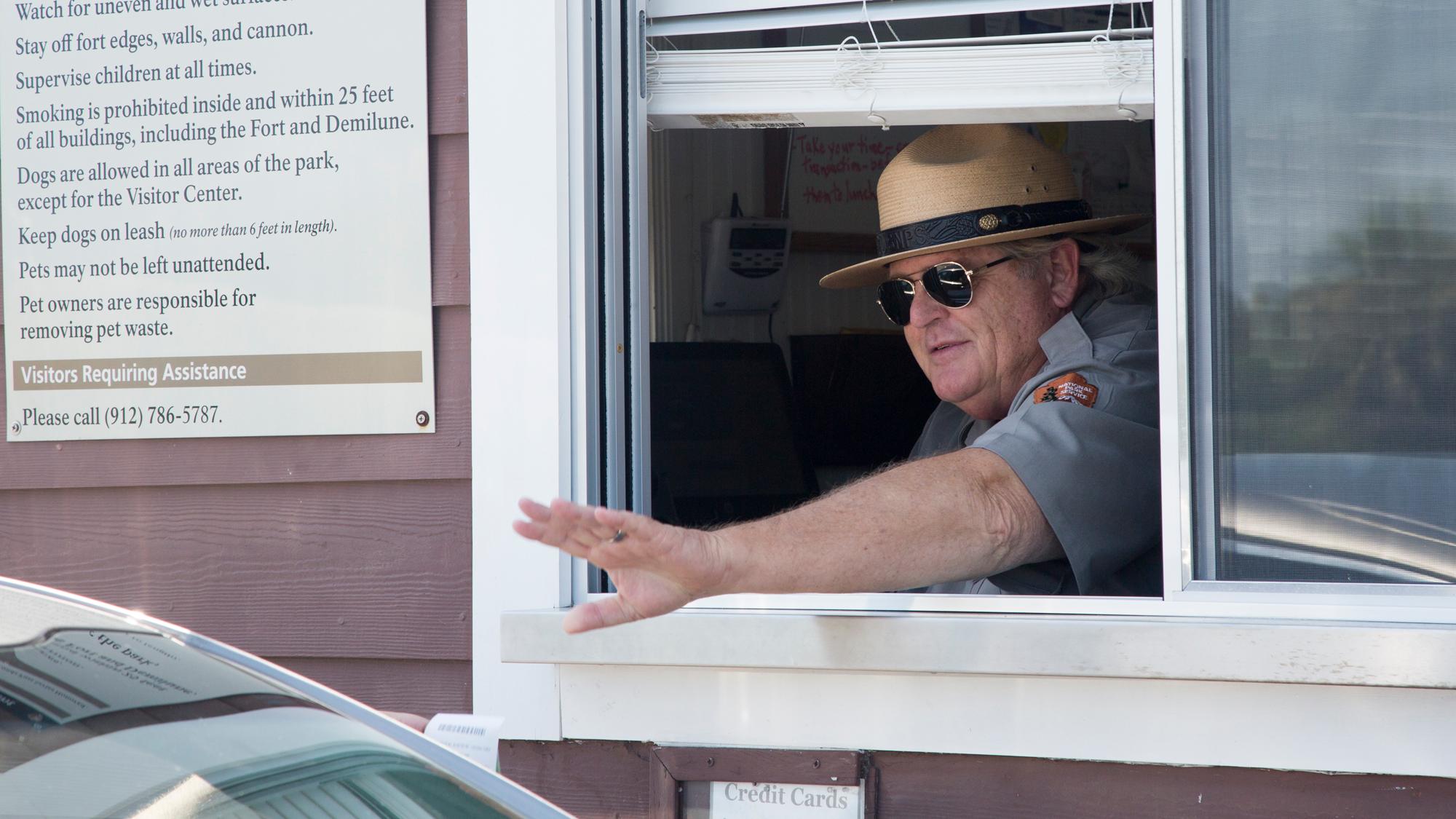 A park ranger, in a window, waving to a car. 