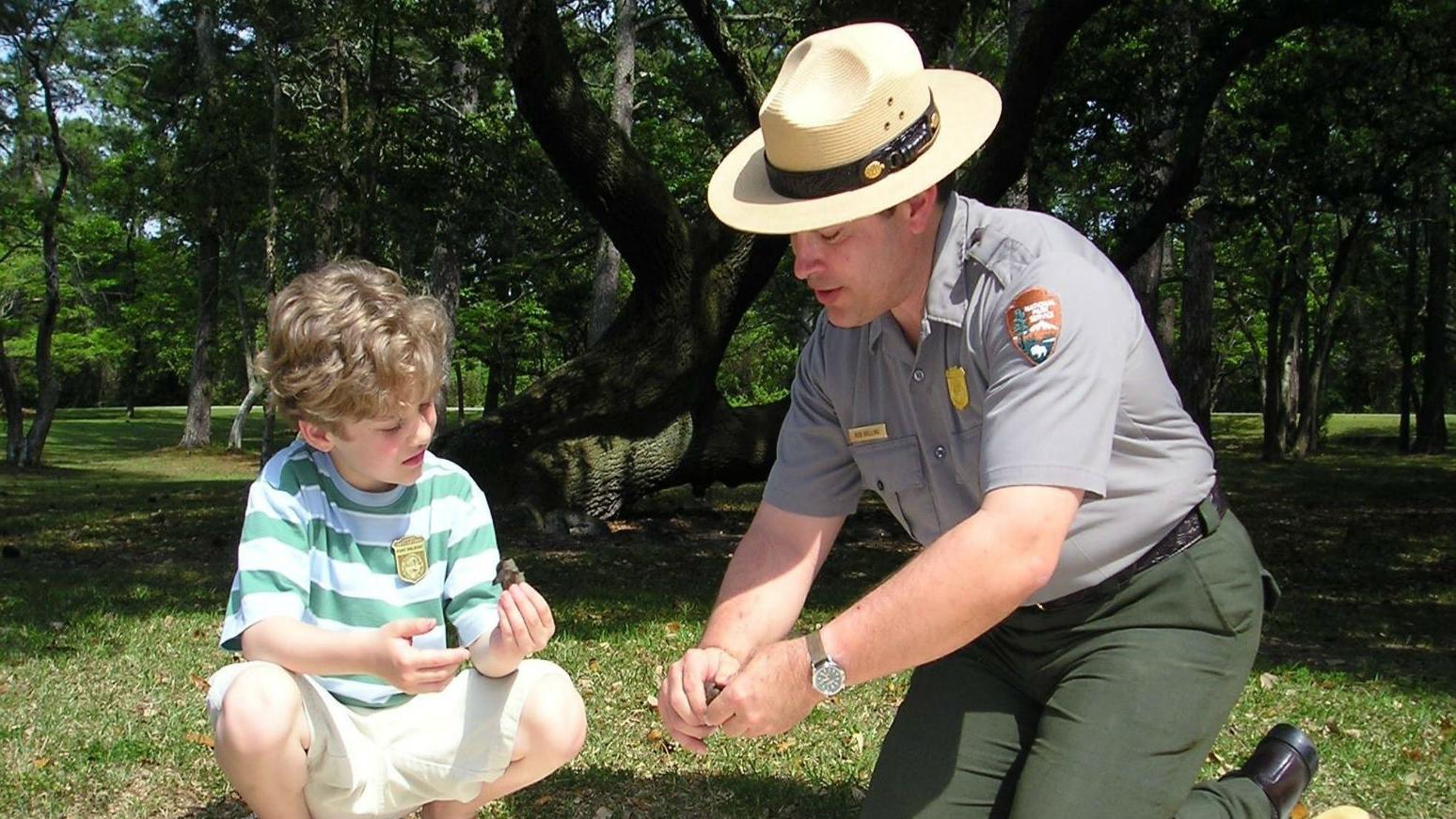 color photo of a ranger at left kneeling showing tree bark to a young boy sitting right