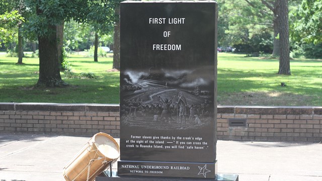 A black stone monument with the words First Light of Freedom. Behind is a stone wall and grass field