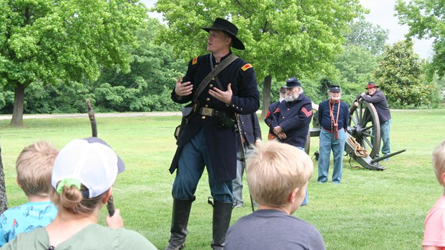 A man wearing a blue Civil War uniform and hat speaking to a crowd in front of a cannon.