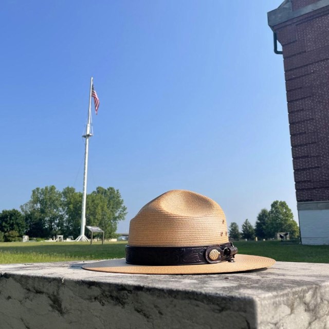 A straw park ranger flat hat with a leather band sits on concrete in front of a tall American flag.