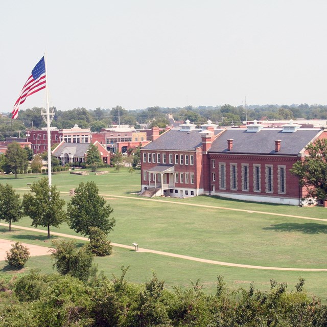 An aerial photo of the grassy parade ground with a tall flagpole and red brick visitor center.