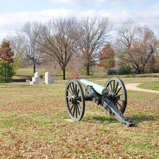 A cannon sitting in a grassy field in the fall, with bare trees and orange leaves on the ground.