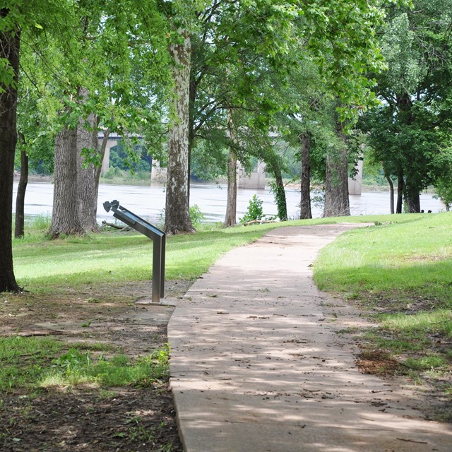 A paved walkway with green grass and trees on either side. A cantilever wayside panel to the left.