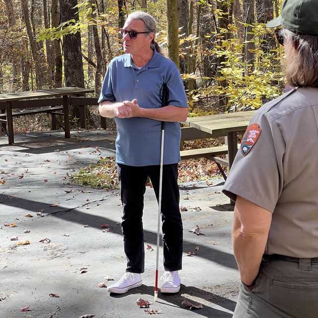 A person wearing sunglasses and holding a white-tipped cane surrounded by trees.