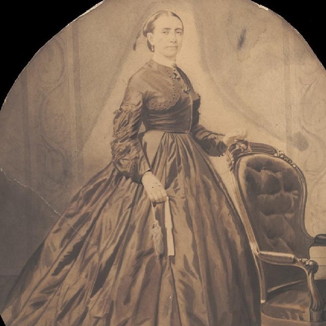 A portrait of a woman in a dark colored dress, looking at the camera with her hand on a chair