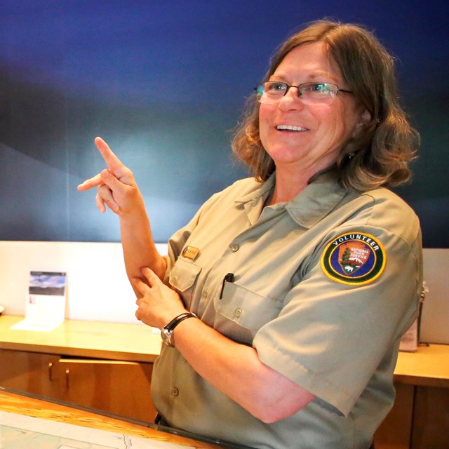 A person in an NPS volunteer uniform makes the letter K in American Sign Language with their hands.