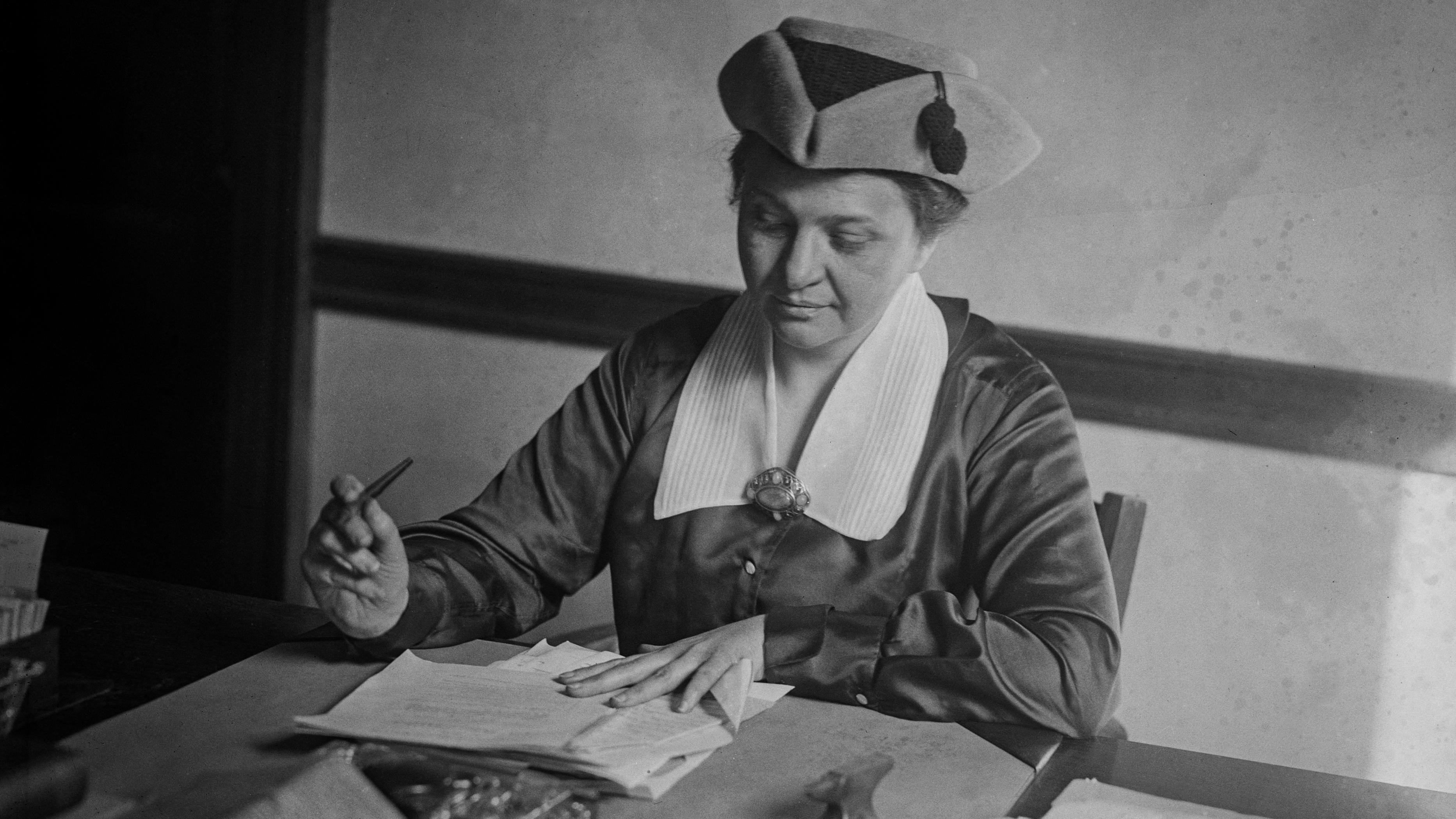 Frances Perkins sitting at a desk, holding a pen and looking down at an open notebook.
