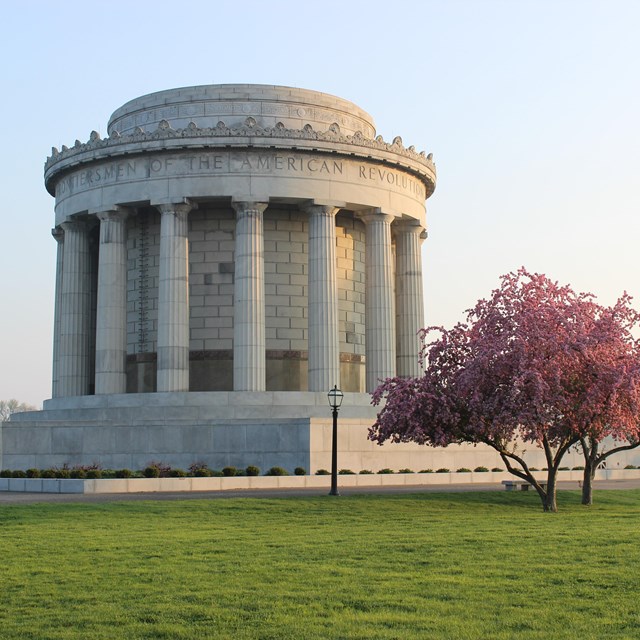 A large round gray colonnaded building with a flowering Redbud tree and green lawn in the foreground