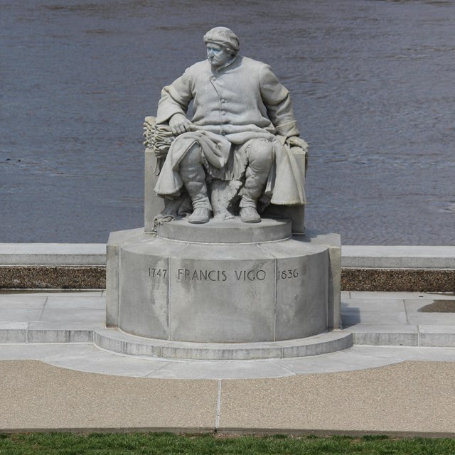 Gray granite statue of Francis Vigo in a long coat sitting on a bench with his arm on stacked furs
