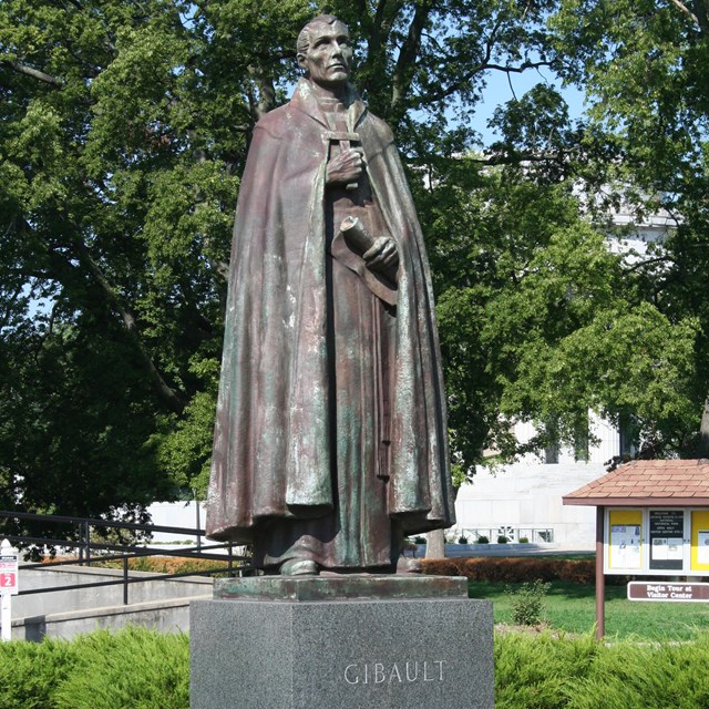 Bronze statue of Pierre Gibault wearing priests robes and a cloak, holding a cross.