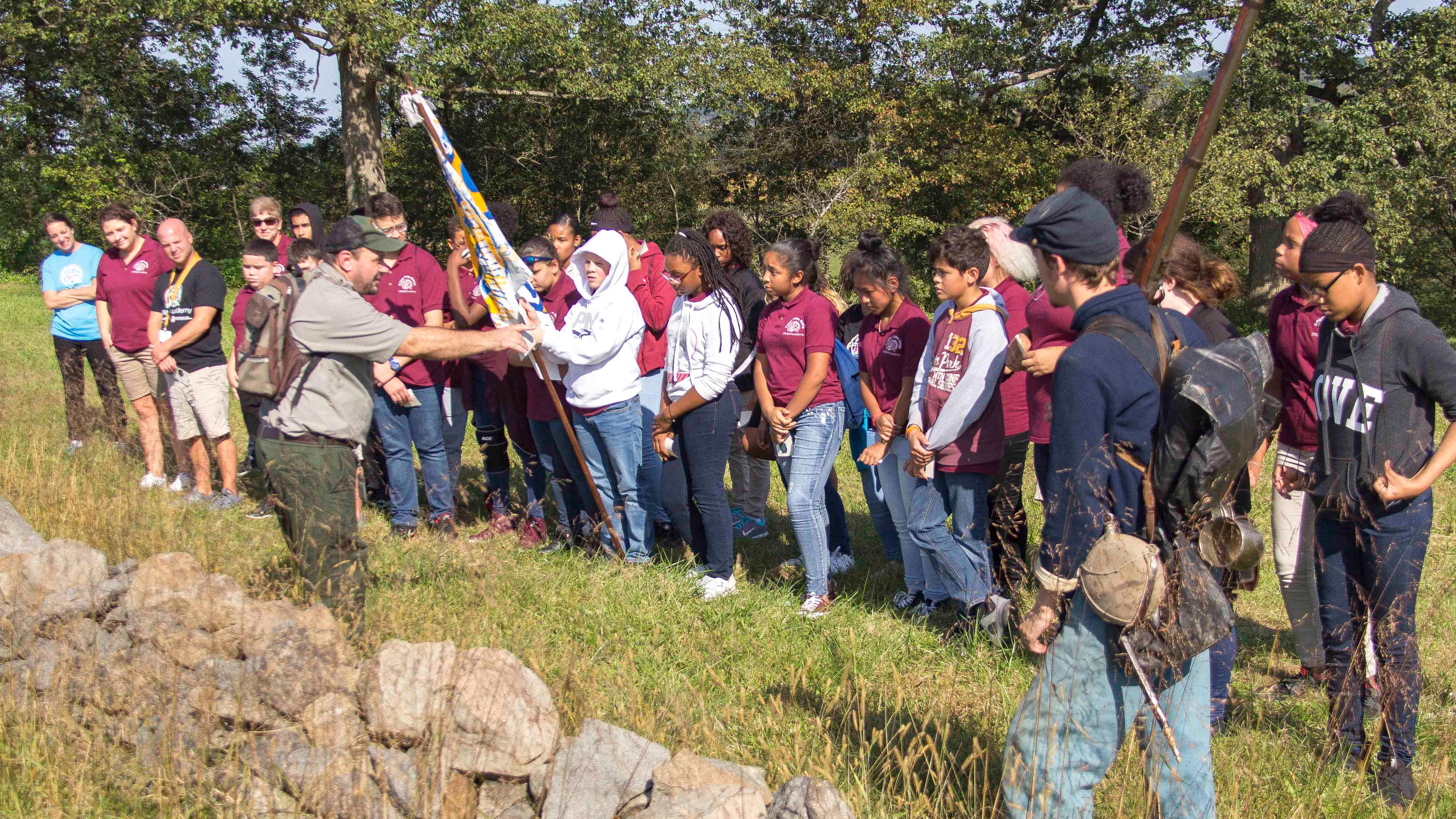 A park ranger leads a group of children on an education program along a stone wall with flags. 