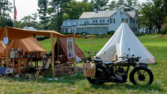 A World War II era motorcycles sits next to a brown tent. A white tent and white house are behind it