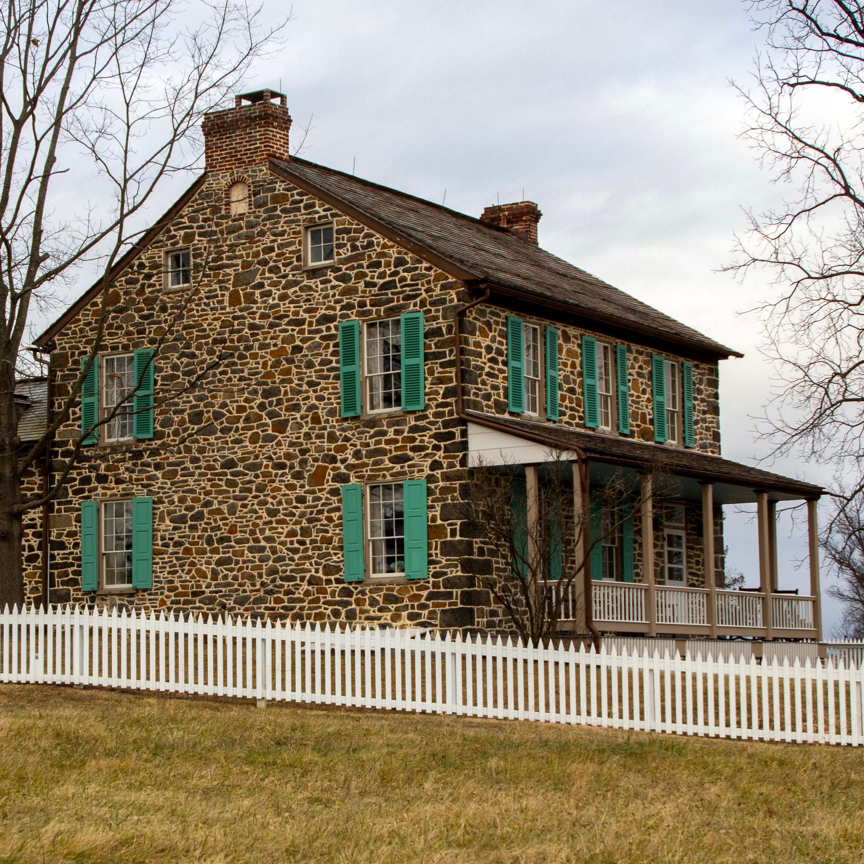 A two-story stone house with green shutters and front porch on the right has a white picket fence.