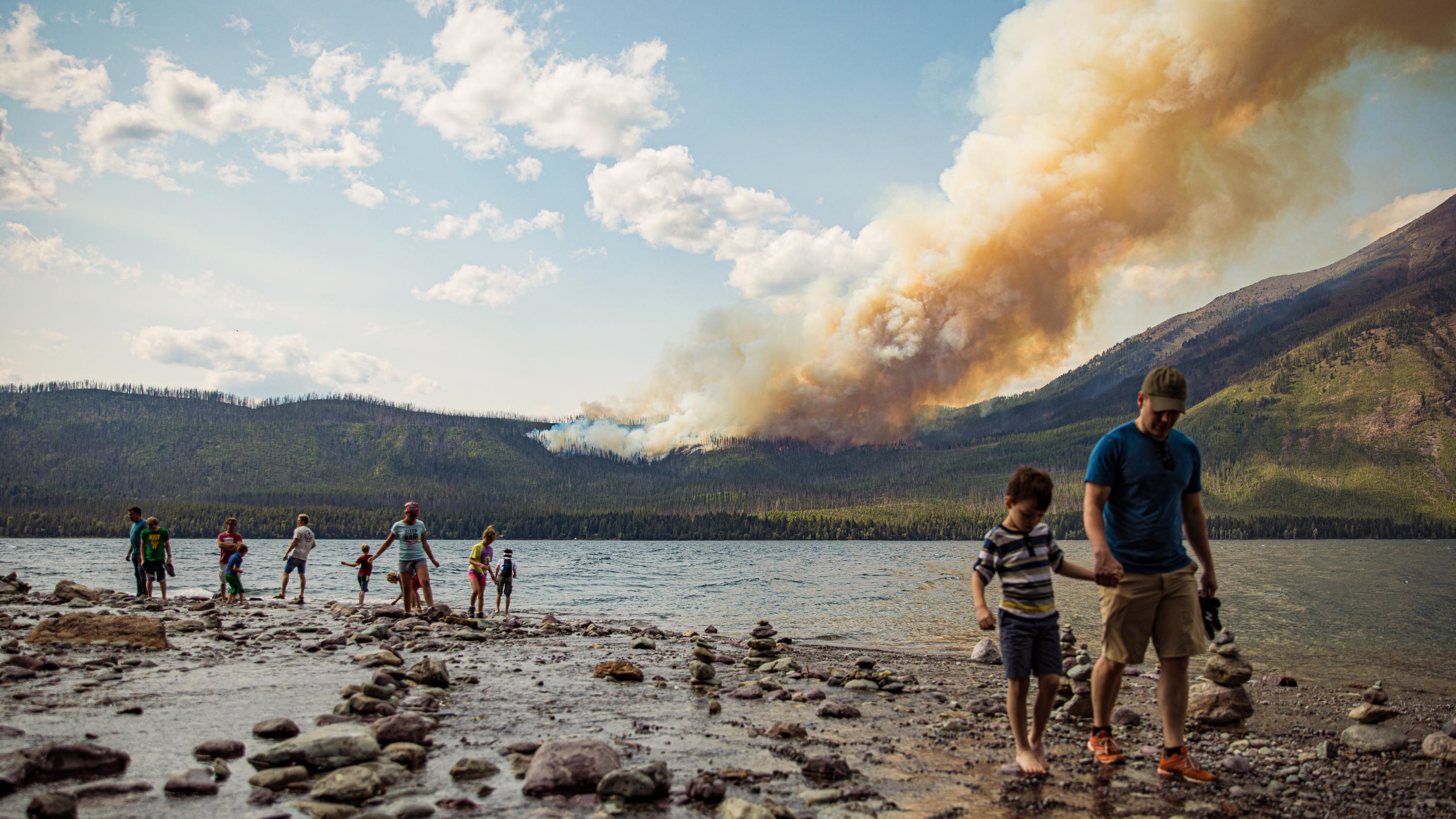 People walk on a beach with a wildfire behind them.