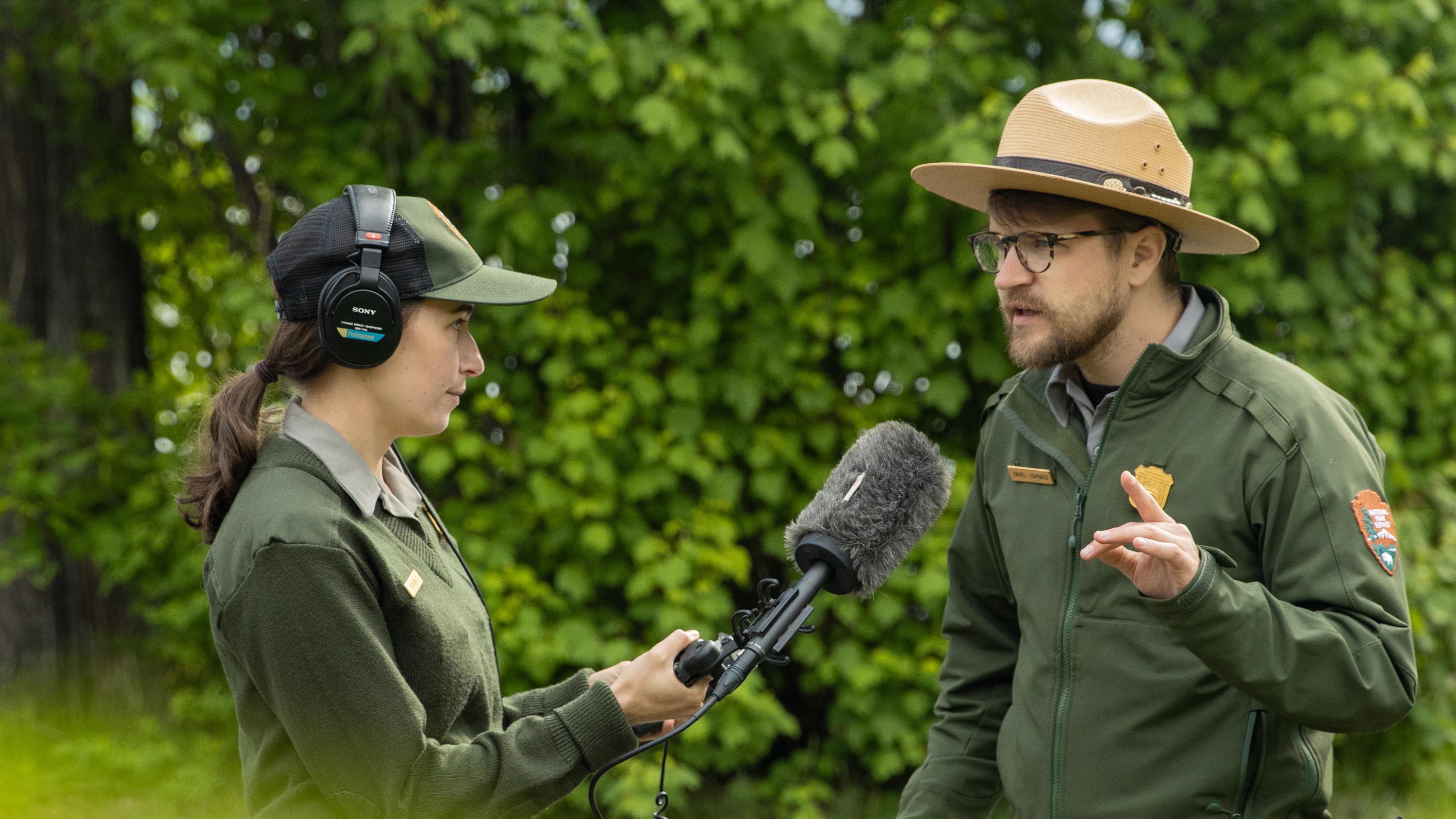 Two park rangers talk outdoors with a microphone between them.