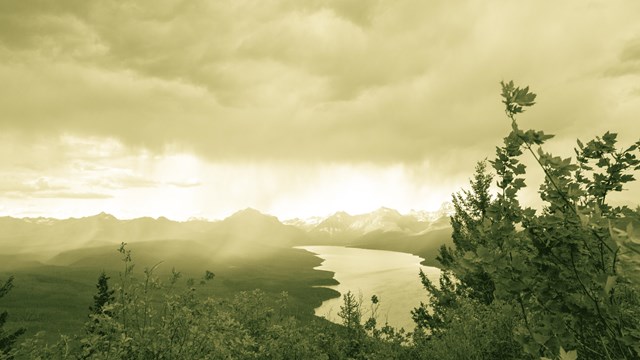 A bolt of sunlight shines through cloudy and hazy skies, hitting mountains and a lake below.
