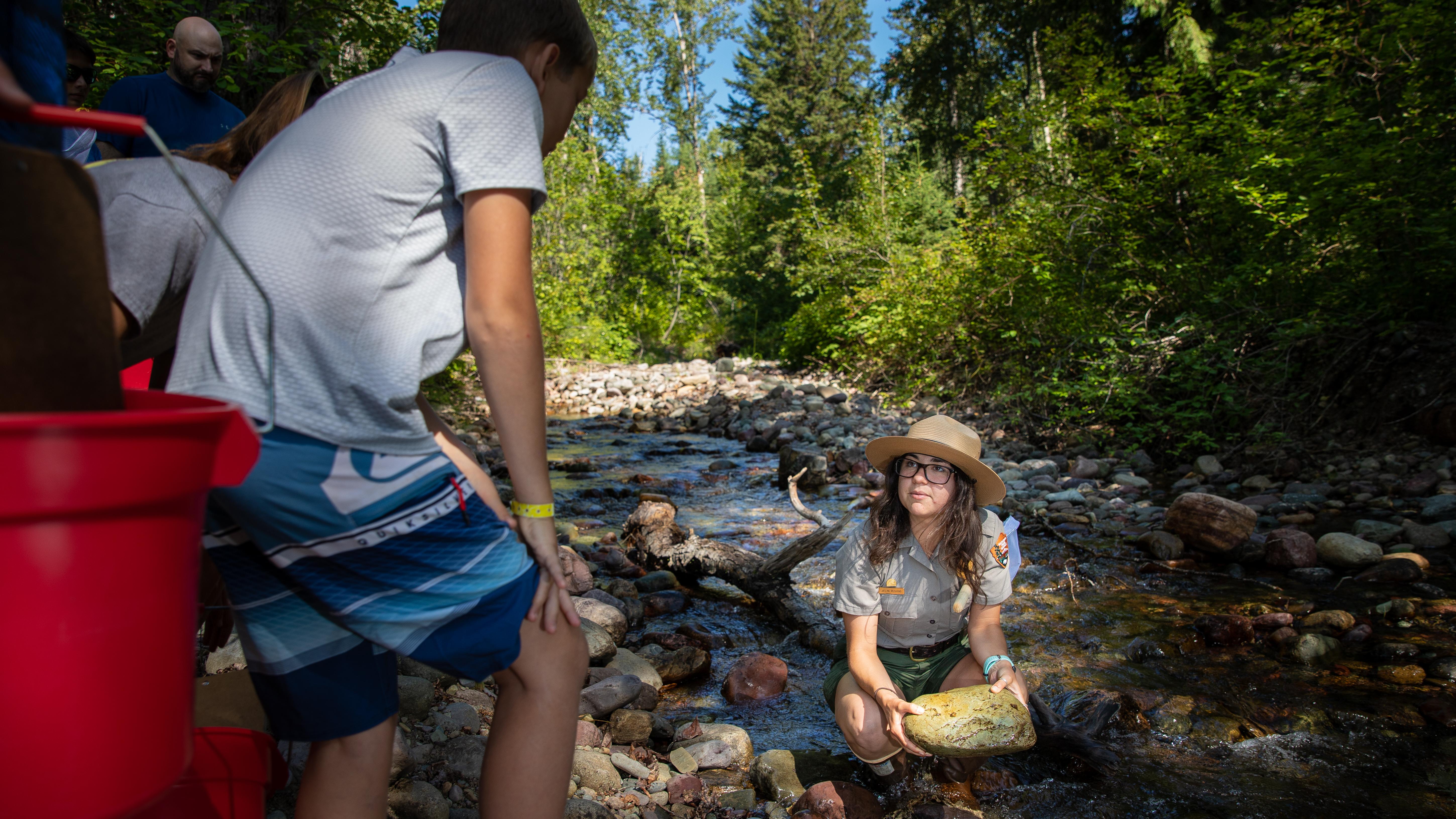 A ranger squats in a creek holding a large yellow rock while talking to a group of kids on the bank.