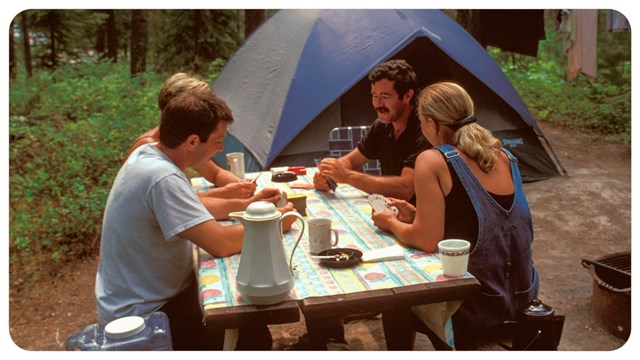 Group of people sit by a tent. Trees in the background. 