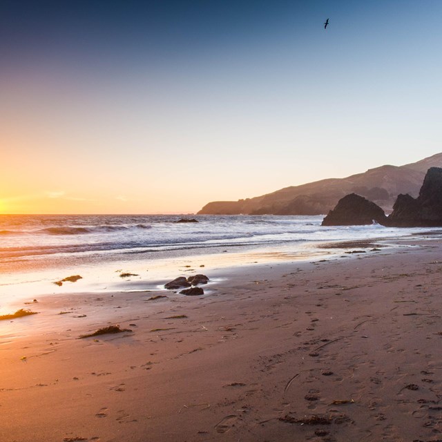 The sun sets on a beach with large rock formations. Short buildings are visible in the background.