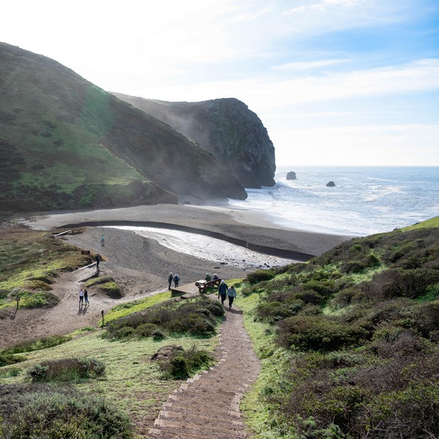 Hikers walk down a path toward a cove draped in fog on a sunny day.