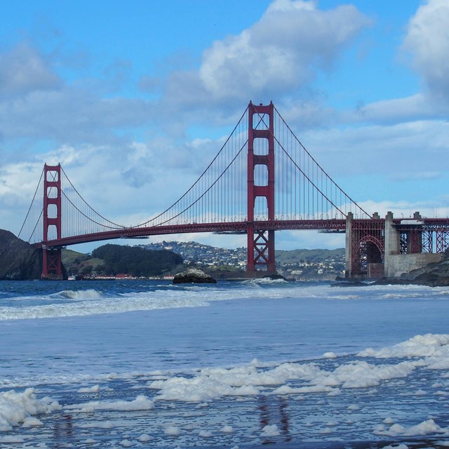 Sea foam covers a beach with fort point and the golden gate bridge in the background.