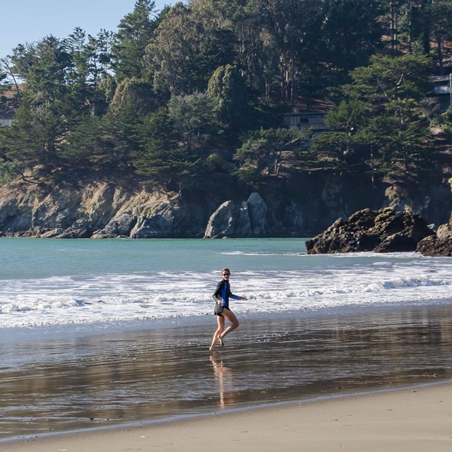 A person runs along the beach with two large rocks surrounded by trees and mountains