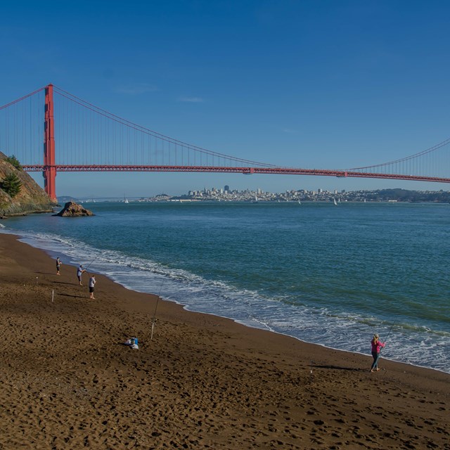 People fish and walk on the beach with the Golden Gate Bridge in the background.