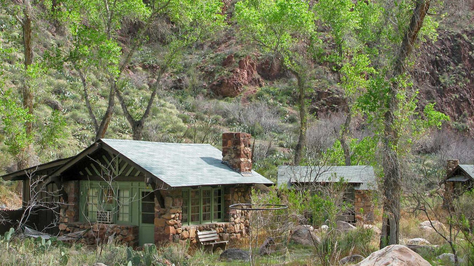 Several rustic stone and wood cabins underneath green cottonwood trees and canyon walls. 
