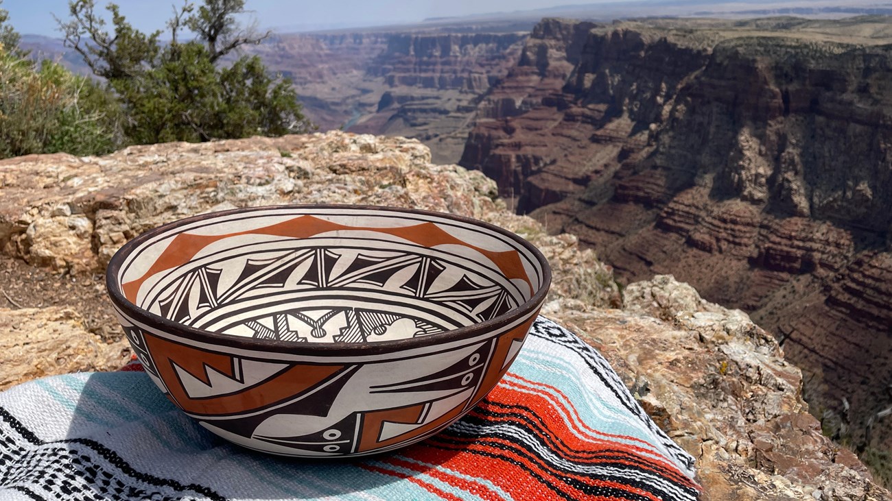 A geometric red, black, and white ceramic bowl sits on a colorful blanket with a deep canyon behind.