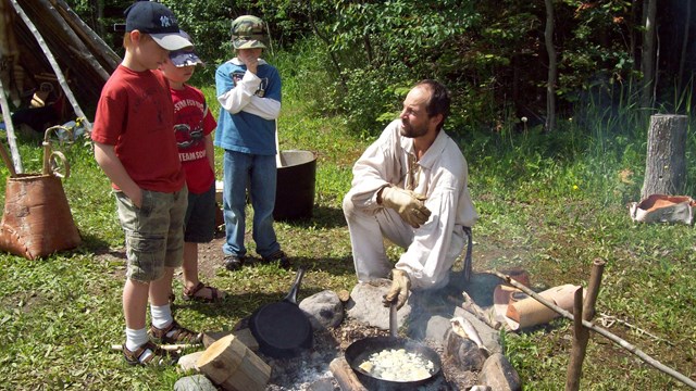 Three kids watch a costumed interpreter cook over an outdoor fire.