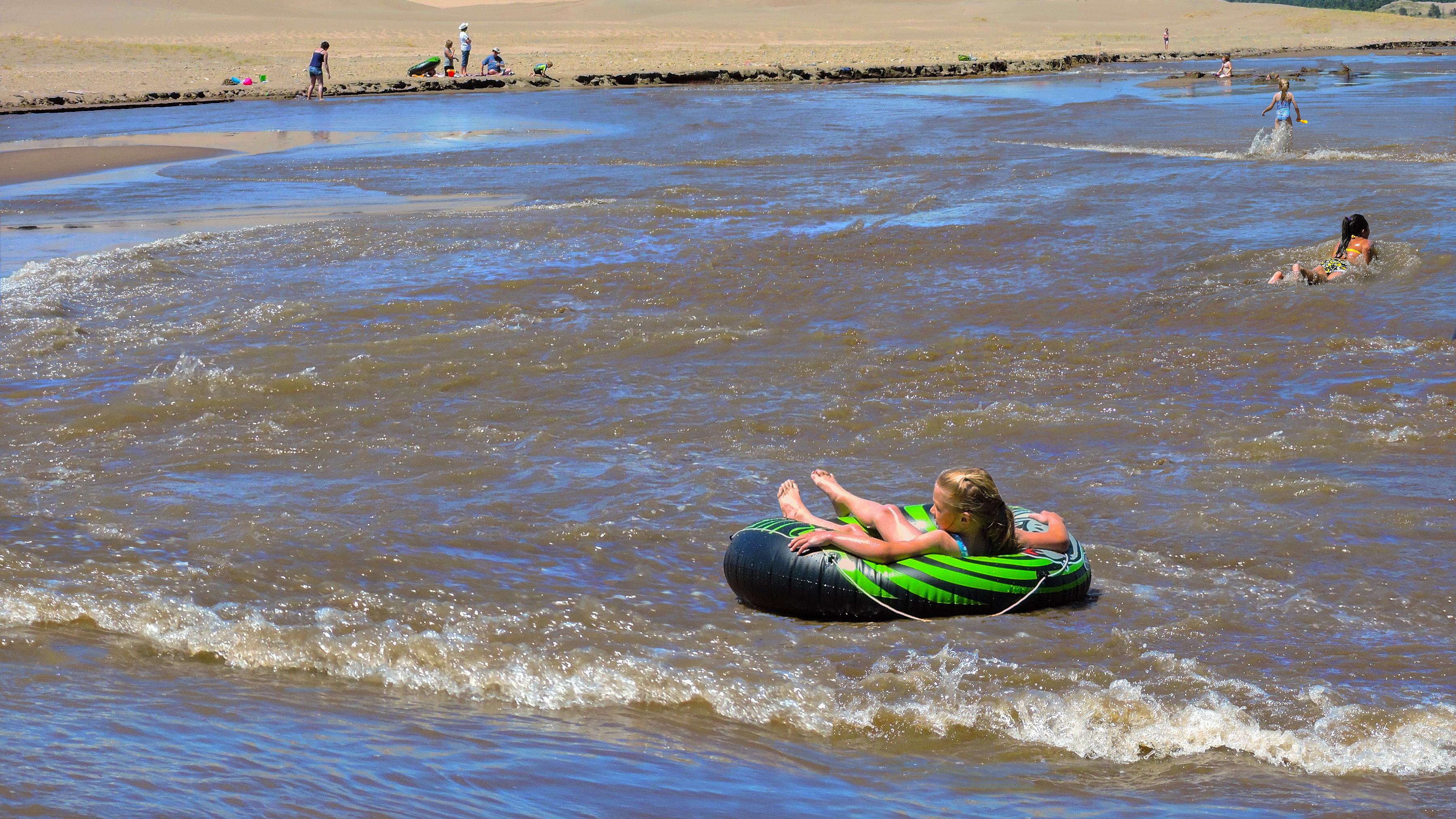 A girl floats on a tube on a wave of Medano Creek in front of dunes and a mountain
