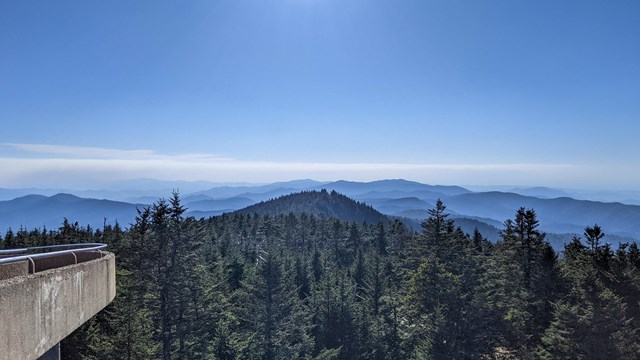 A sweeping mountain view with evergreen trees in the foreground. Left frame shows a paved walkway.