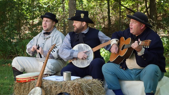 Three men sitting on hay bales playing music instruments. 