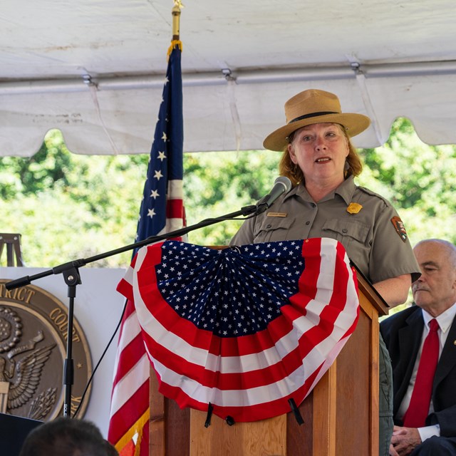 A woman in a ranger uniform stands at a lectern with bunting.