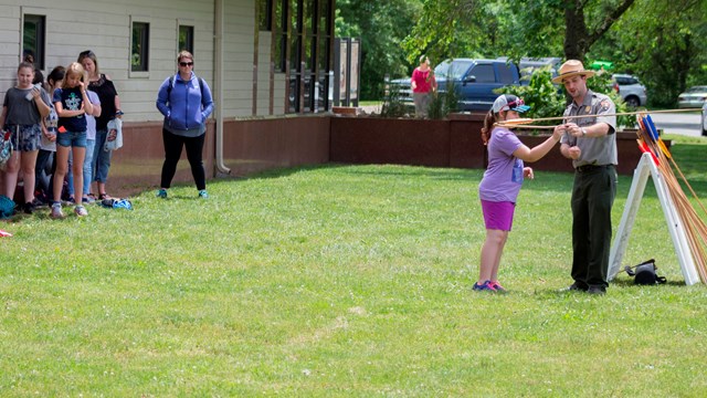 A ranger in a gray shirt and tan flat hat helps a school student hold onto a spear thrower.