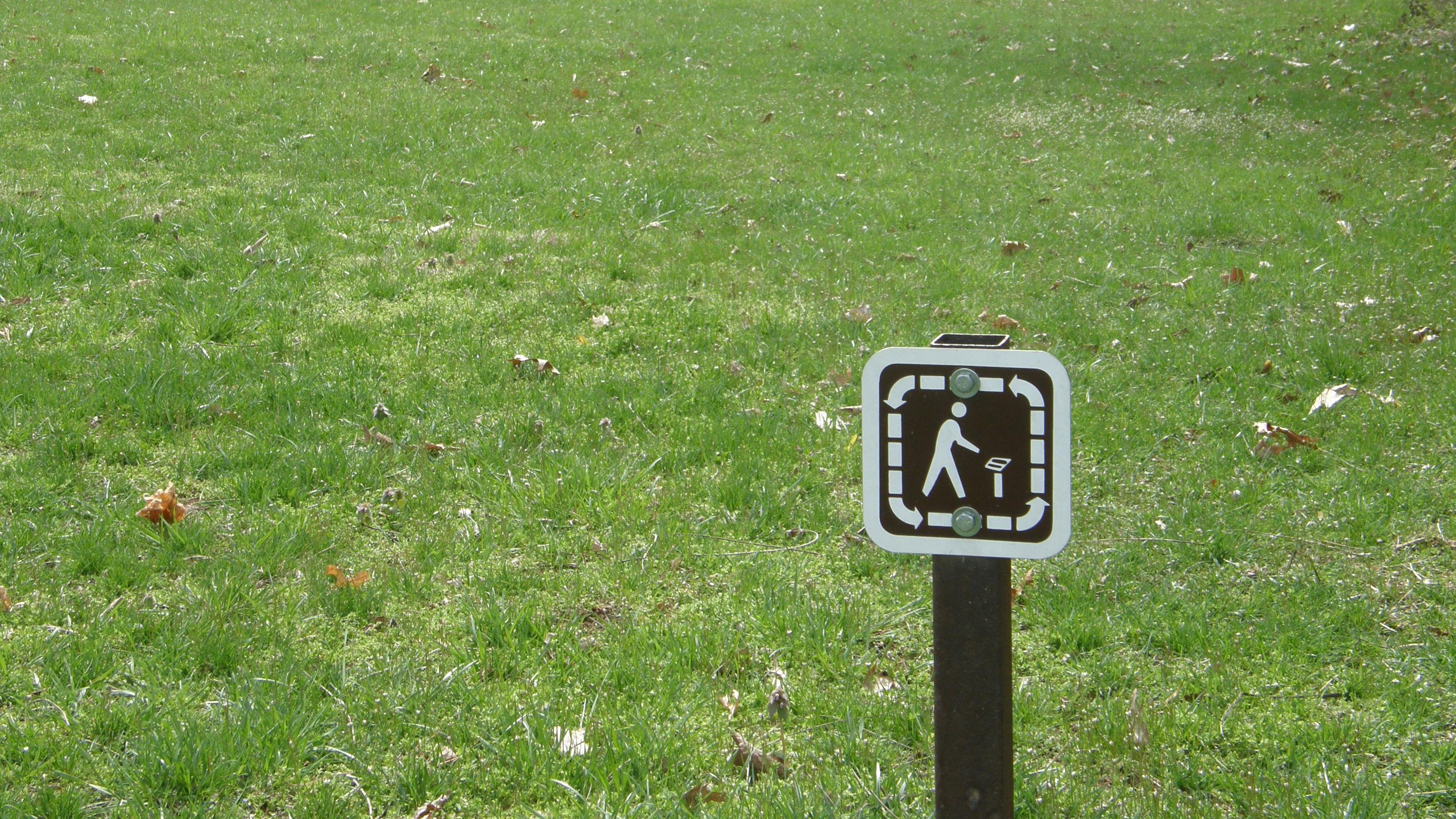 A small brown trail sign in front of a wide area of grass on a trail