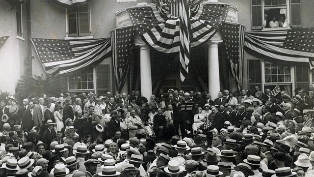 A large group of people crowd around the front of a house draped in American flags.