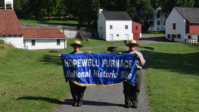 Two park rangers hold a Hopewell Furnace banner in the historic village.