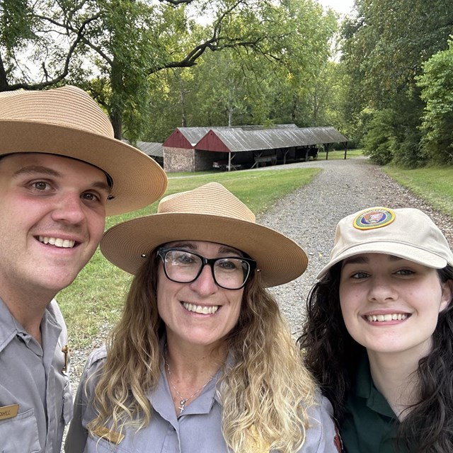 Three people smile outside. One is wearing a volunteer uniform hat, the other two are park rangers. 