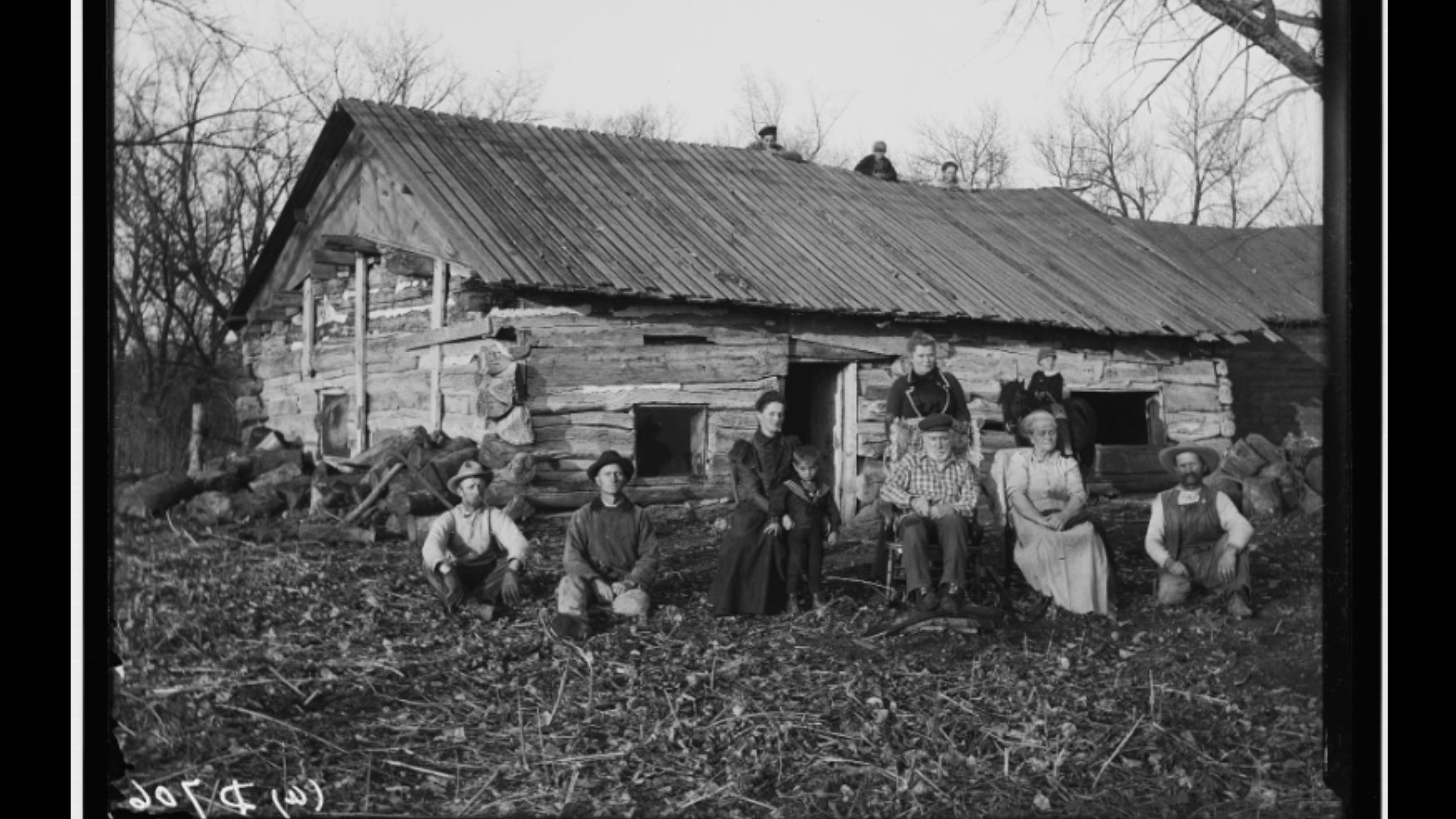 Black and white photo, white family outside log home.