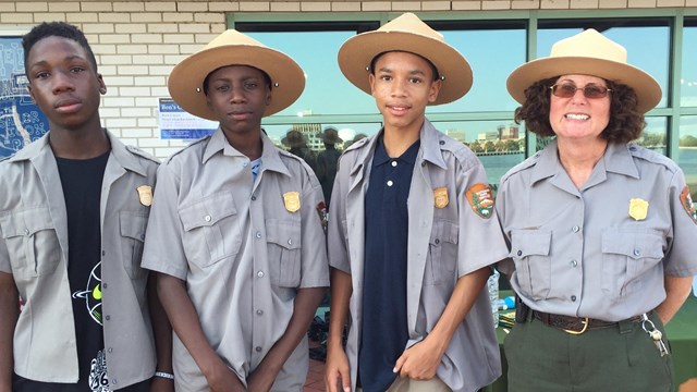 Color image of three teen boys wearing ranger shirts and hats while standing next to a ranger.