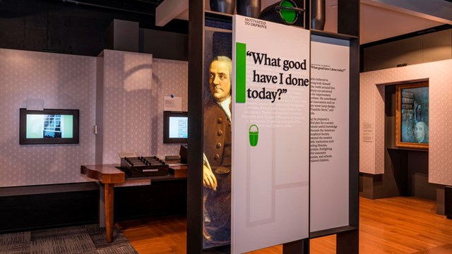 Color photo of an exhibit panel with an image of Benjamin Franklin, and exhibits behind.