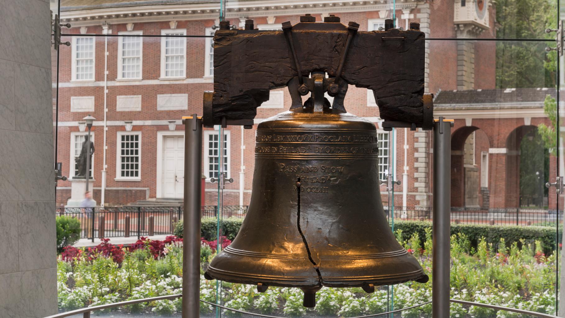 Color photo of the Liberty Bell with Independence Hall visible through the glass behind it.
