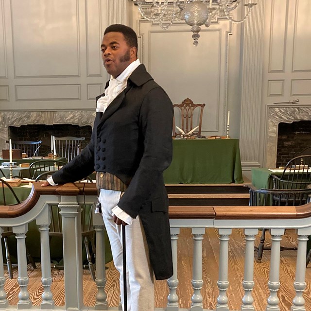 A man in clothing from the 1800s stands in the Assembly Room of Independence Hall.