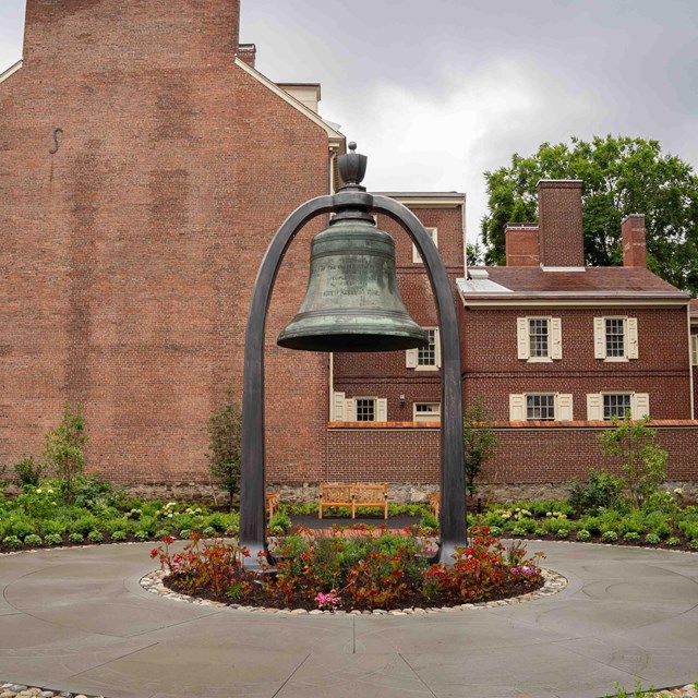Color photo of garden with a large bell suspended from metal arch. 