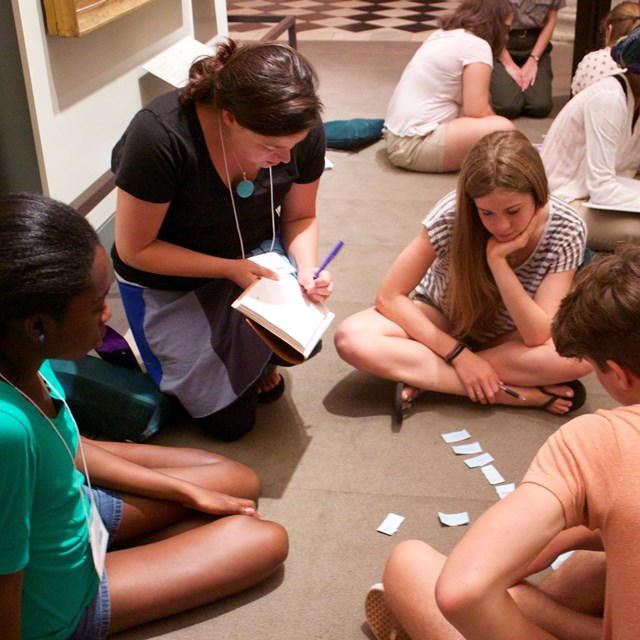 Color photo of a teacher sitting on the floor surrounded by a group of teens.